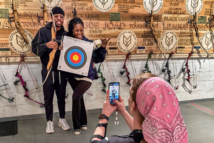 Two smiling people holding archery target with person taking photo, bows on wall in background.