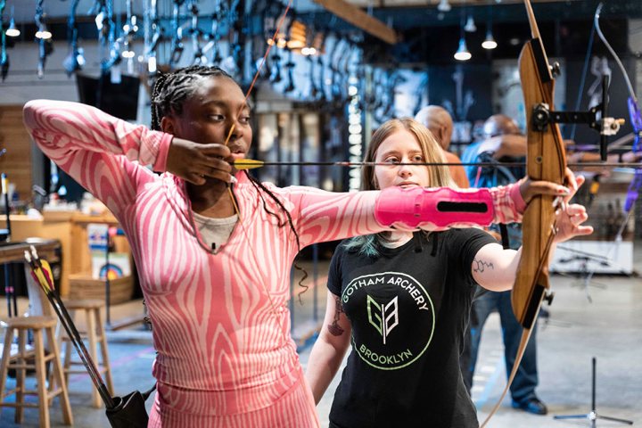 Woman in pink aiming a bow, guided by an instructor in a busy archery range.
