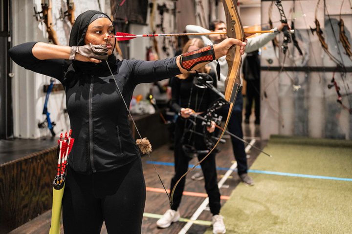 Person in black archery gear aims bow and arrow in indoor range.