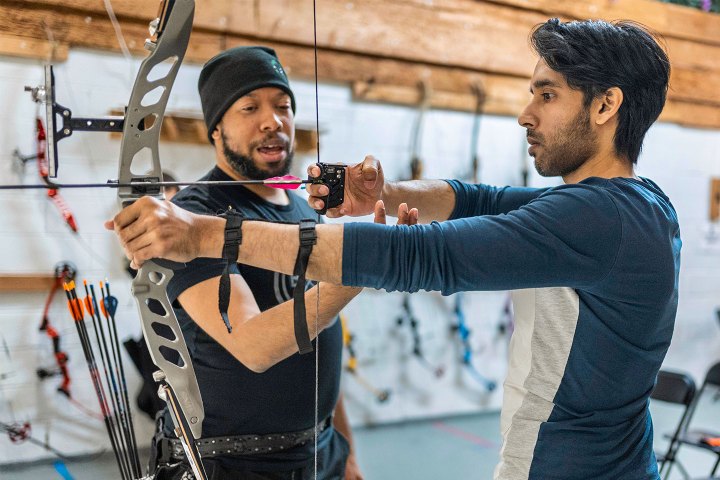 Two men practicing archery indoors, one instructing the other.