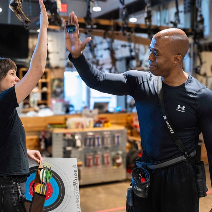 Two people high-fiving in an archery range, holding bows, with targets in the background.